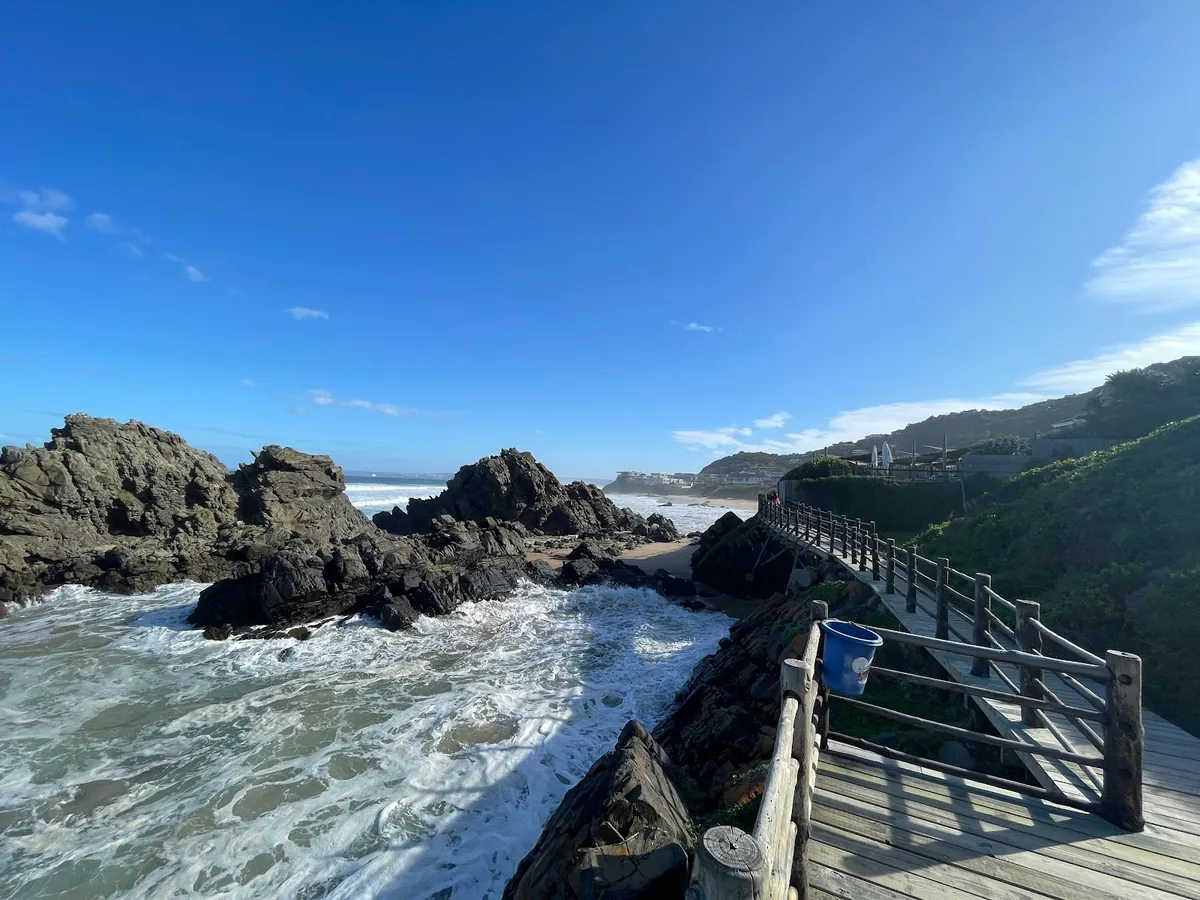 Timber boardwalk bridging rocks at Keurboomstrand, whitewater surging underneath, forested hillside homes above — Plettenberg Bay
