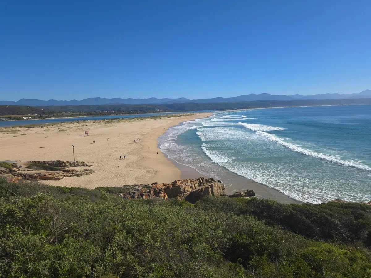 Lookout Beach at Plettenberg Bay from an elevated viewpoint — long curving white sand sweeping toward the Keurbooms river mouth with Tsitsikamma mountains in the distance