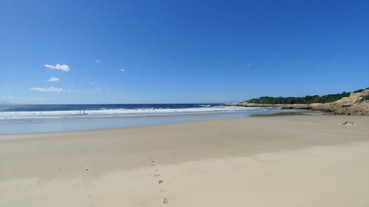 Quiet empty sand at the Robberg end of Lookout Beach with distant surfers and a small rocky headland to the right