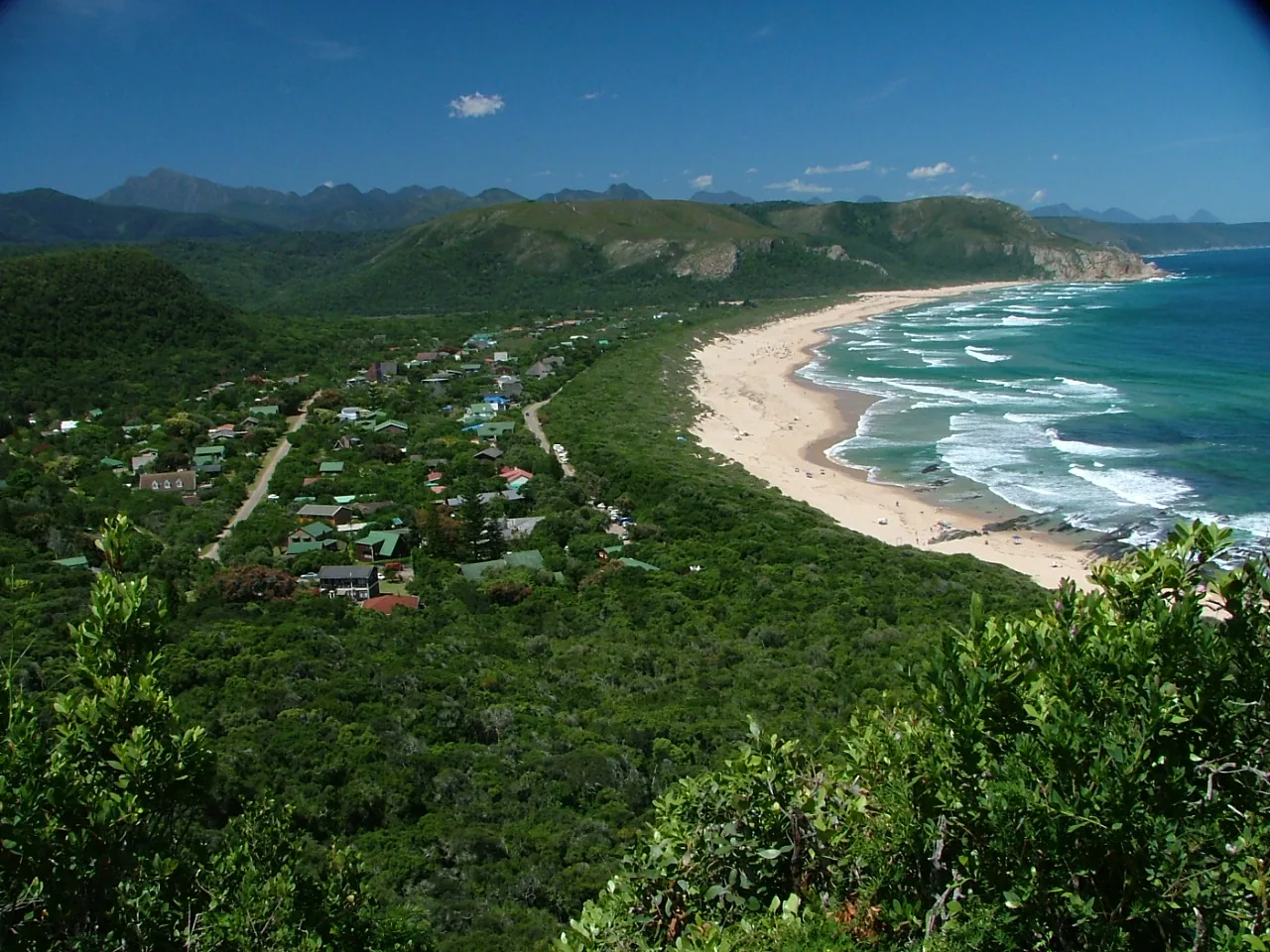 Aerial of Nature's Valley — the village nestled in indigenous forest, long curving white beach, Tsitsikamma mountains on the horizon