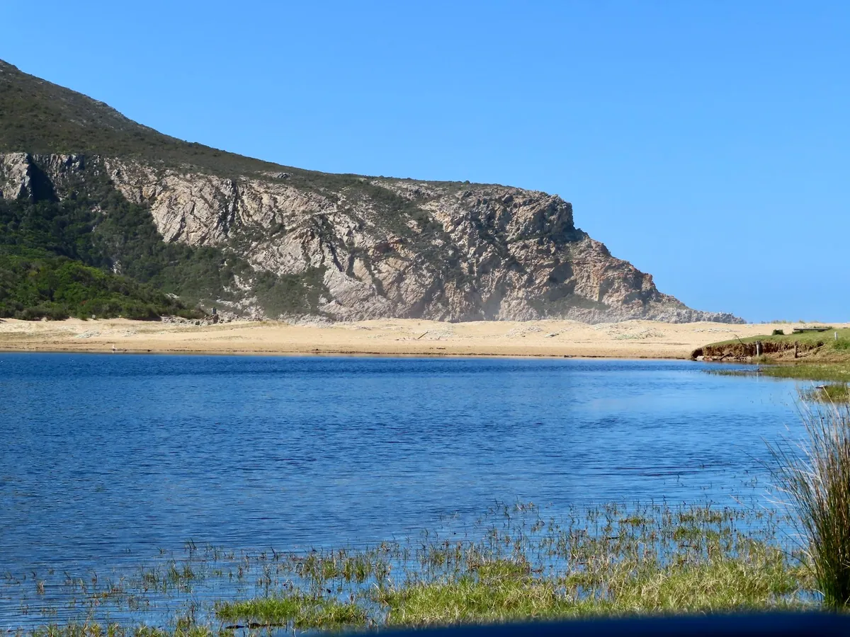 Groot River lagoon at Nature's Valley with a rocky sandstone cliff rising behind the sandbar and reed-fringed shallows in the foreground