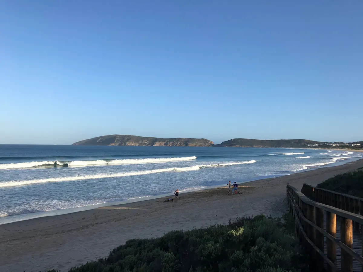 Robberg Beach under clear afternoon light — long parallel surf lines rolling in, Robberg Peninsula on the horizon, a handful of walkers on the wet sand