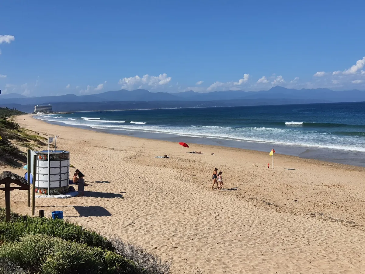 Wide Robberg Beach with a Blue Flag lifeguard flag planted in the sand and the Beacon Isle Hotel visible in the distance