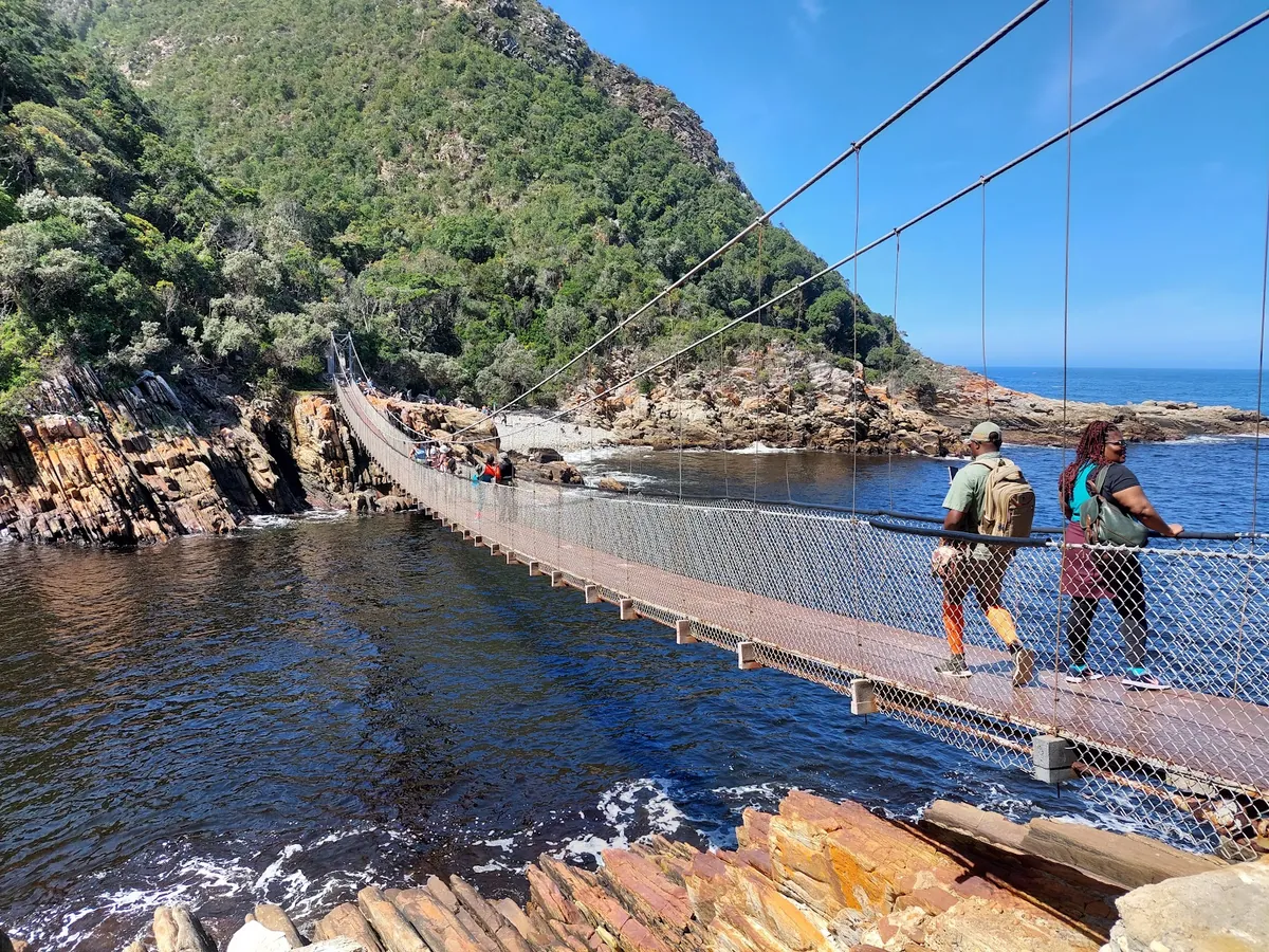 Two walkers mid-crossing on the Storms River suspension bridge in bright sun, fynbos-covered cliffs behind them and the tidal pool visible below