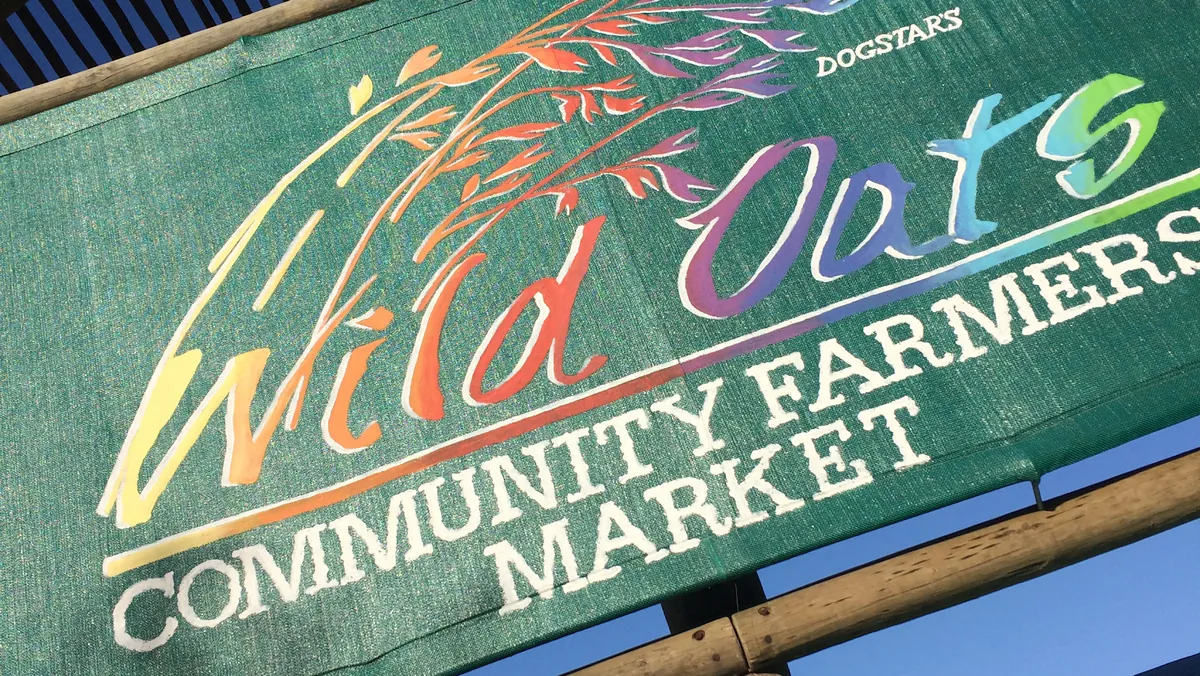Close-up of the painted Wild Oats Community Farmers' Market banner sign — rainbow-coloured hand-painted lettering on green canvas