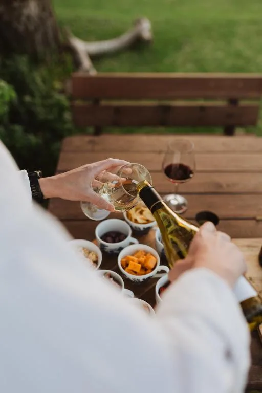 Woman pouring white wine on outdoor wooden deck with snacks and green lawn beyond