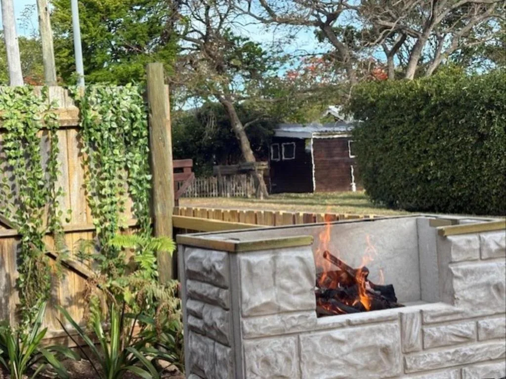 Outdoor fireplace with flames, wooden deck, lush garden, mature trees
