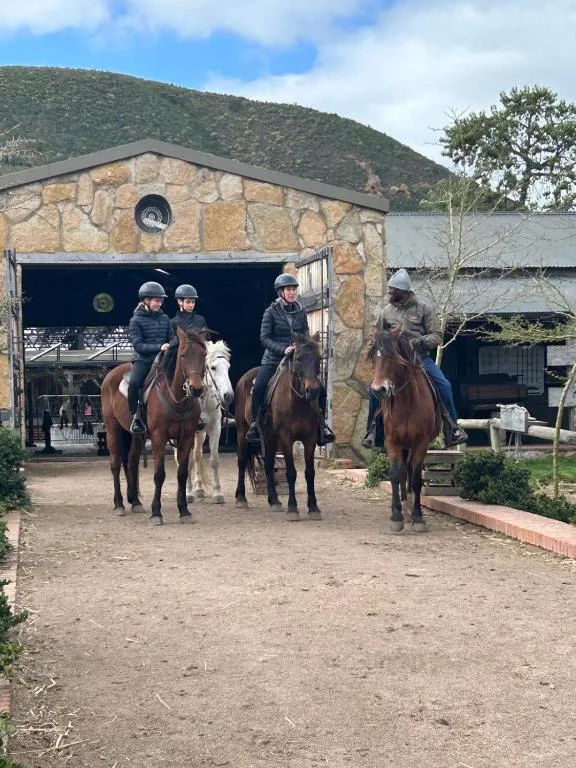 Four riders on horseback outside stone barn building with hillside backdrop