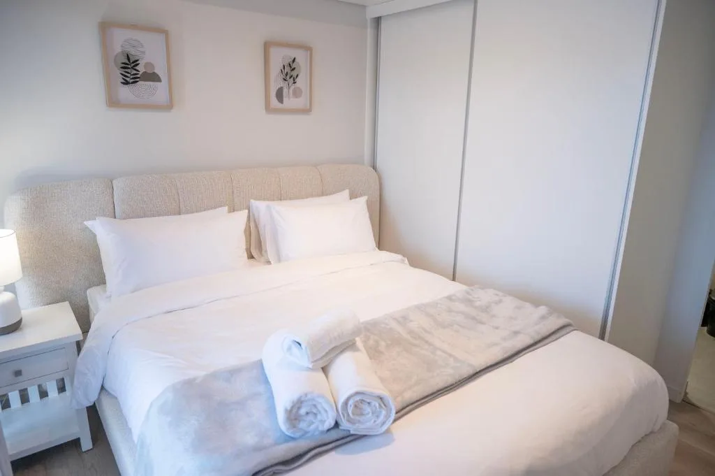 Bright bedroom with queen bed, neutral upholstered headboard, white linens, rolled towels