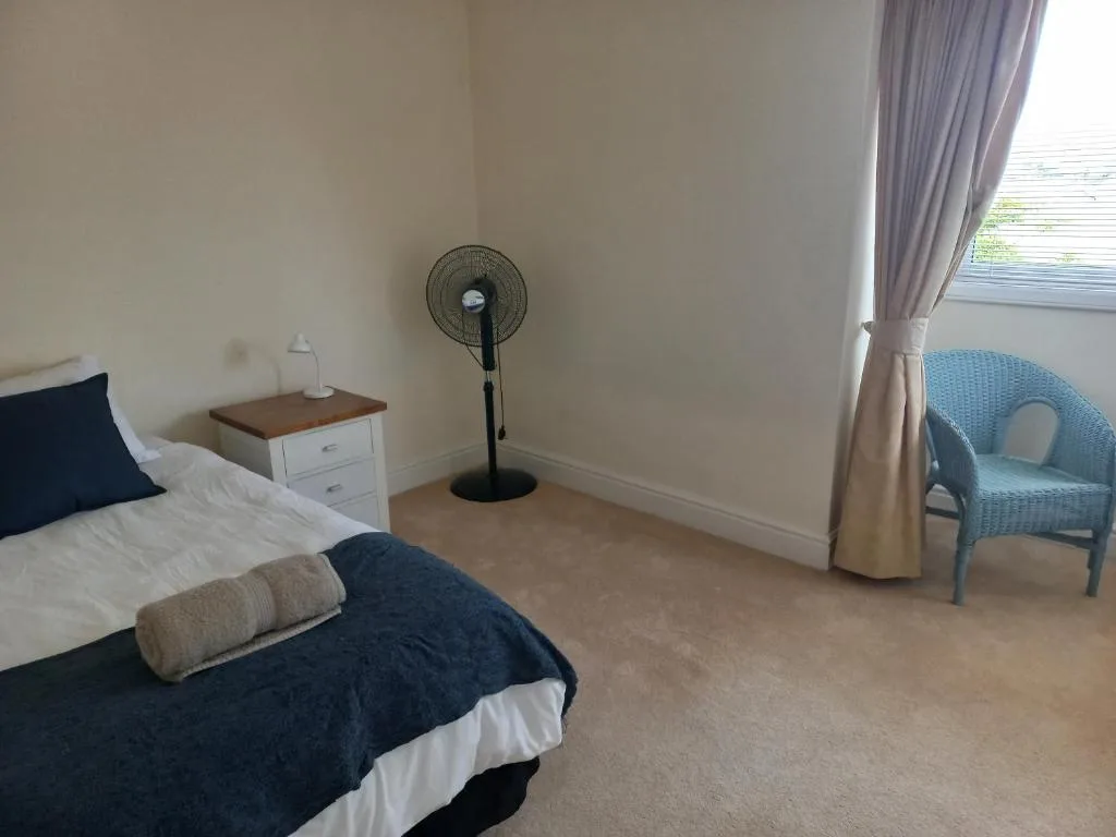 Spacious bedroom with double bed, nightstand, standing fan, and blue chair