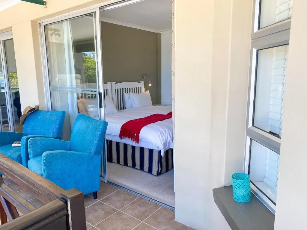 Bright bedroom with striped bed, red throw, and blue accent chairs visible through open sliding doors