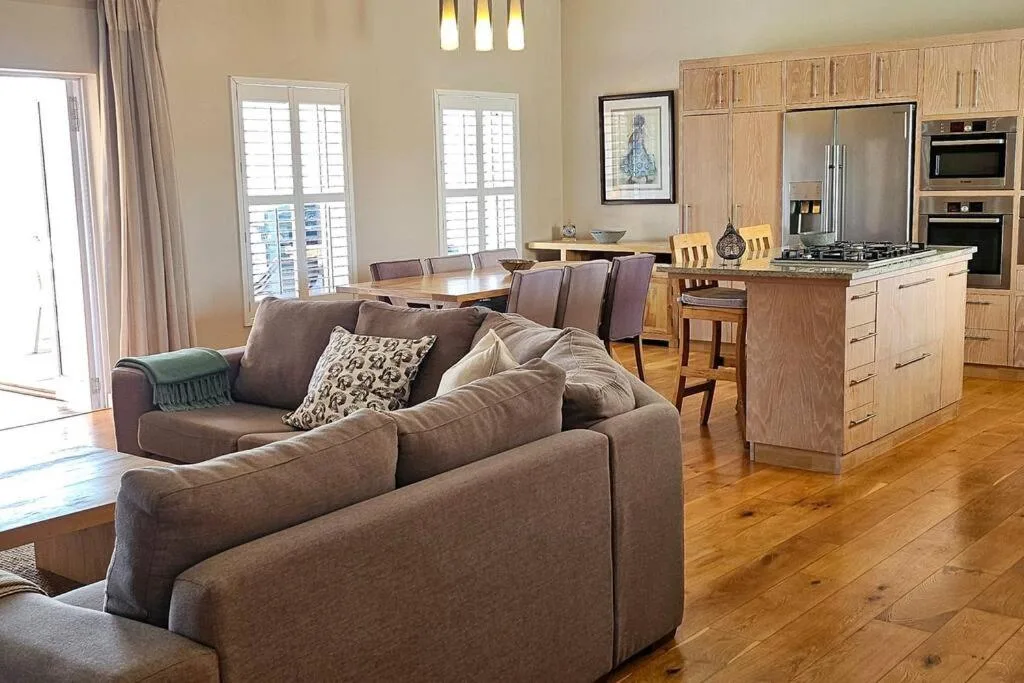 Open-plan lounge with brown sofa, wooden flooring, and kitchen visible behind