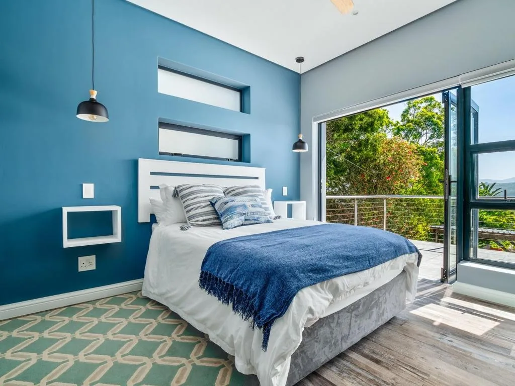 Modern bedroom with teal walls, white bed, and sliding door to garden views