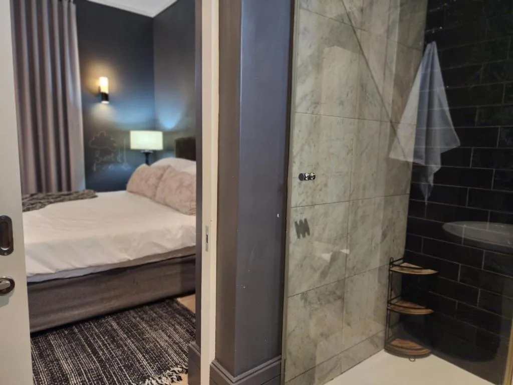 Modern bathroom with grey tile walls and corner shelving, adjoining bedroom view