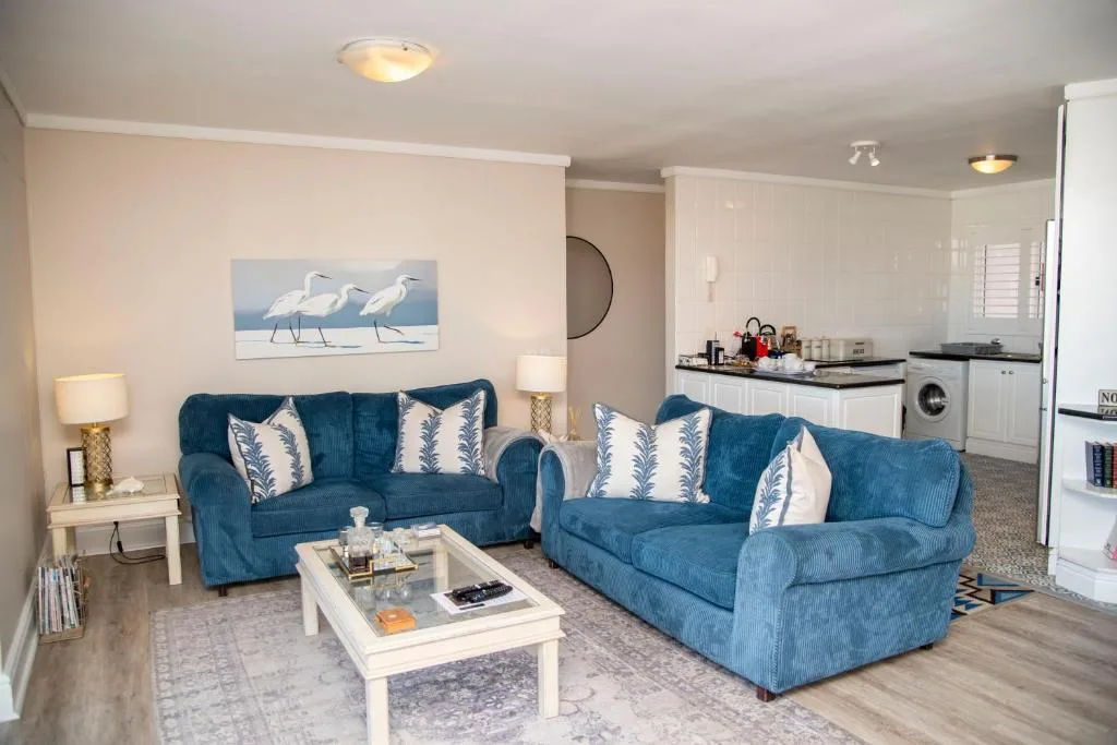 Open-plan lounge with two blue sofas, white coffee table, and kitchen visible