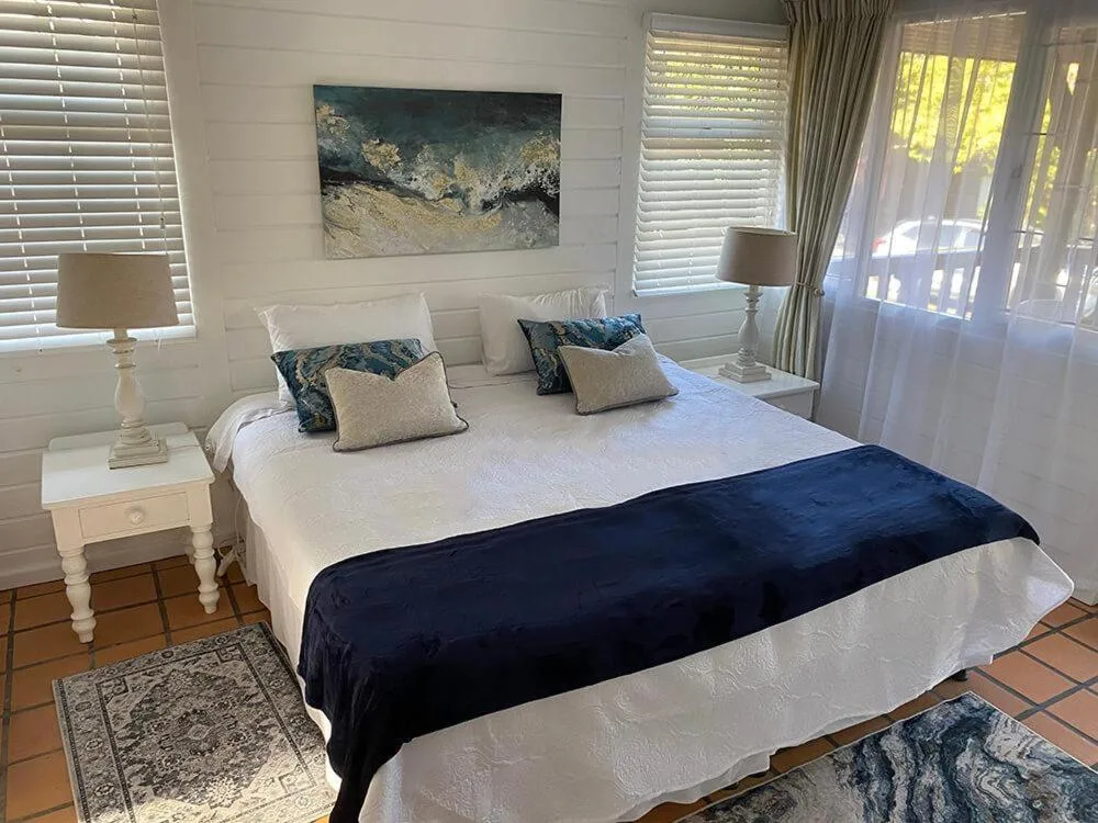 Bright bedroom with queen bed, coastal artwork, white nightstand, shuttered windows
