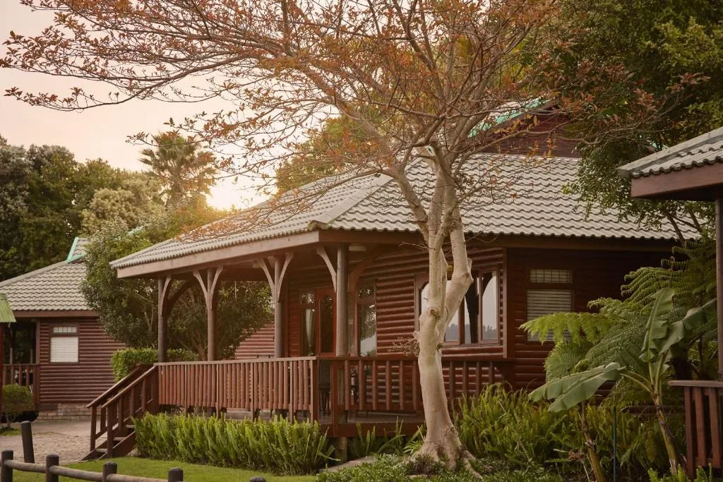 Dark wooden lodge with covered porch, mature tree, lush garden surroundings