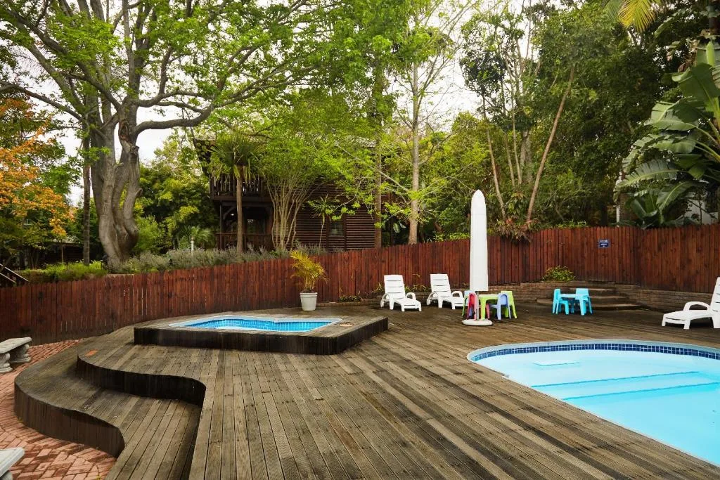 Wooden deck with two pools, loungers, and colorful seating area surrounded by mature trees