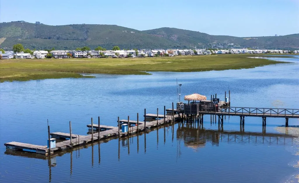 Serene lagoon view with wooden jetty, boats, and Knysna townscape beyond