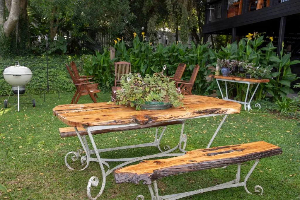 Garden seating area with wooden table, bench, chairs, braai grill, and lush plantings