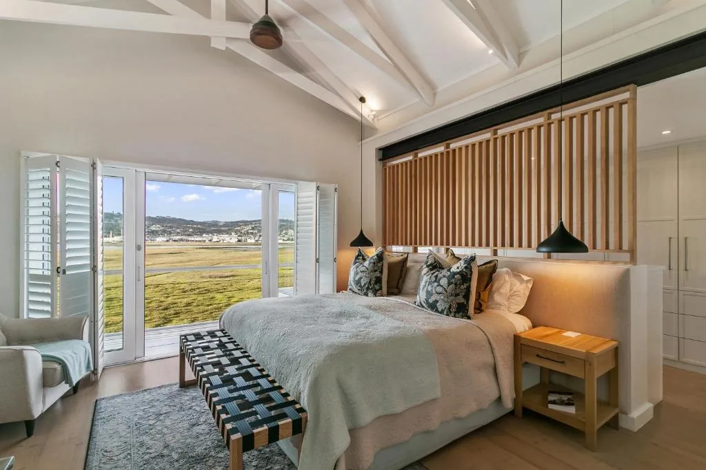 Modern bedroom with bed, exposed beams, and expansive lagoon views through glass doors