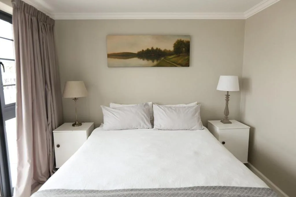 Spacious bedroom with white bedding, bedside tables, and landscape artwork