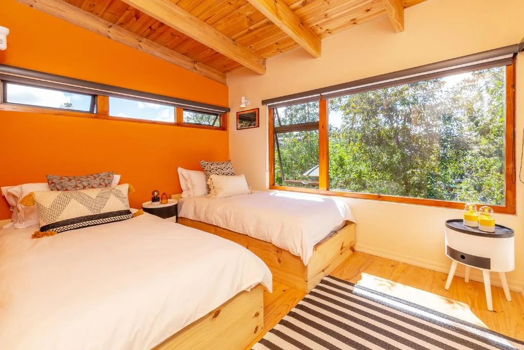 Bright bedroom with two beds, orange accent wall, wooden beams, and large windows