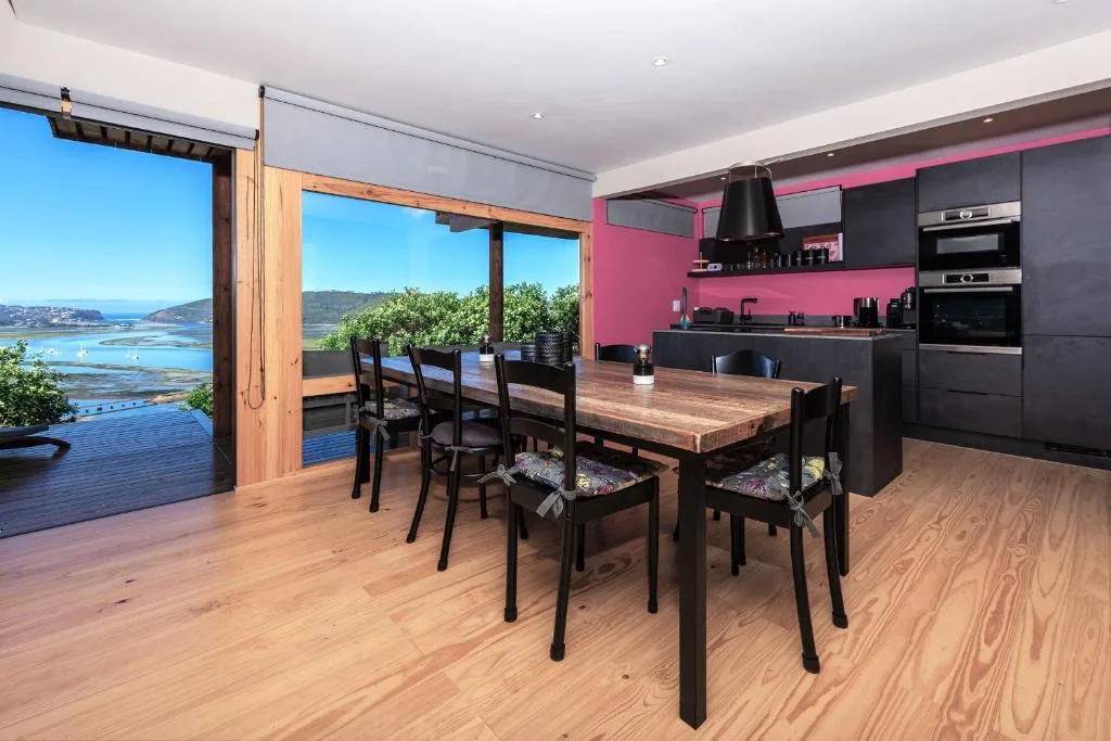 Modern dining room with wooden table, black chairs, pink kitchen, lagoon views