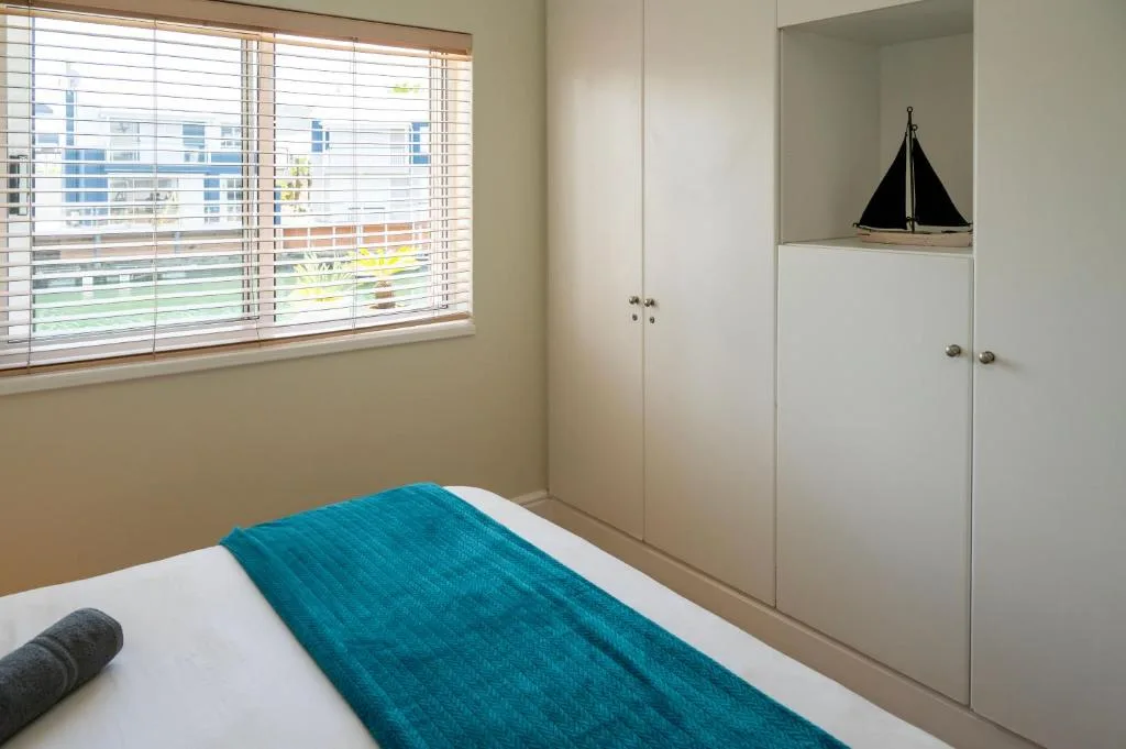 Bright bedroom with teal bedding, built-in wardrobes, and water views through blinds