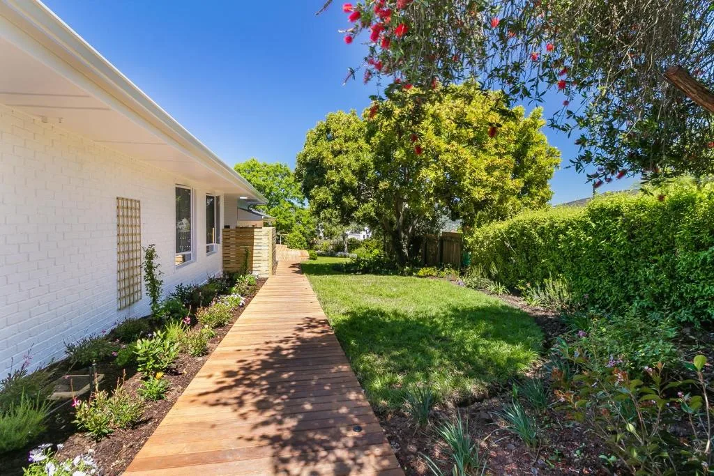 White brick home with wooden pathway through mature garden landscaping