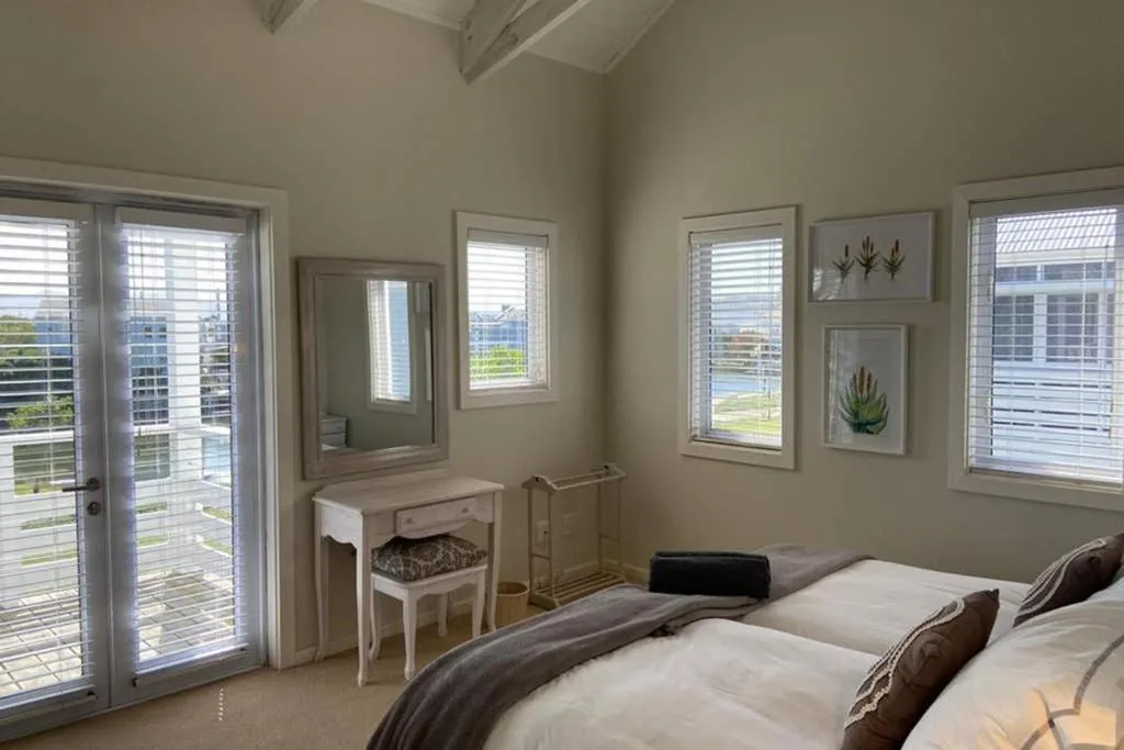 Bright bedroom with twin beds, vanity desk, and louvered shutters overlooking water