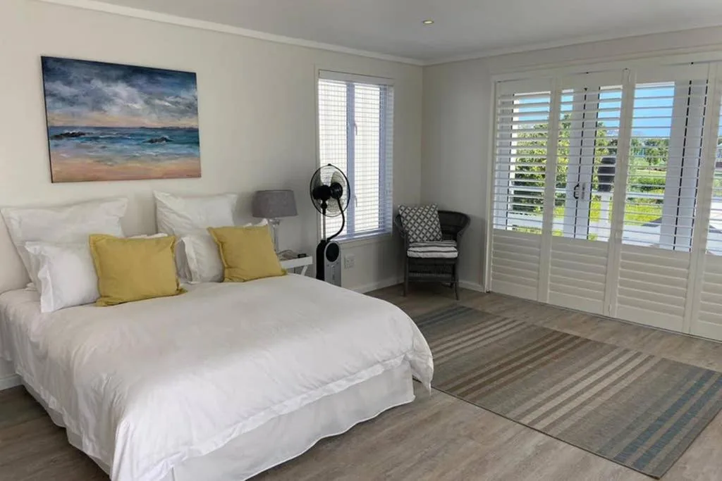 Bright bedroom with white bedding, yellow cushions, ocean artwork, and shuttered doors