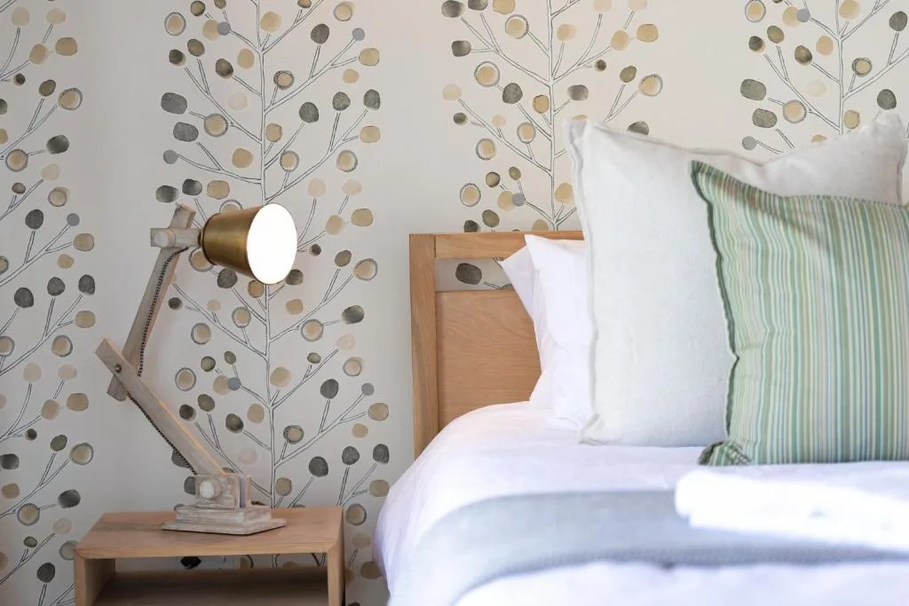 Cozy bedroom with patterned wallpaper, wooden headboard, white pillows, and brass reading lamp