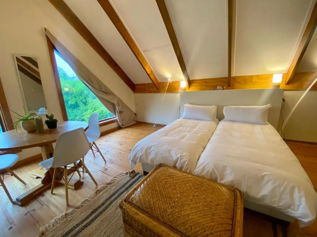 Bright loft bedroom with exposed beams, desk, and triangular window overlooking green landscape