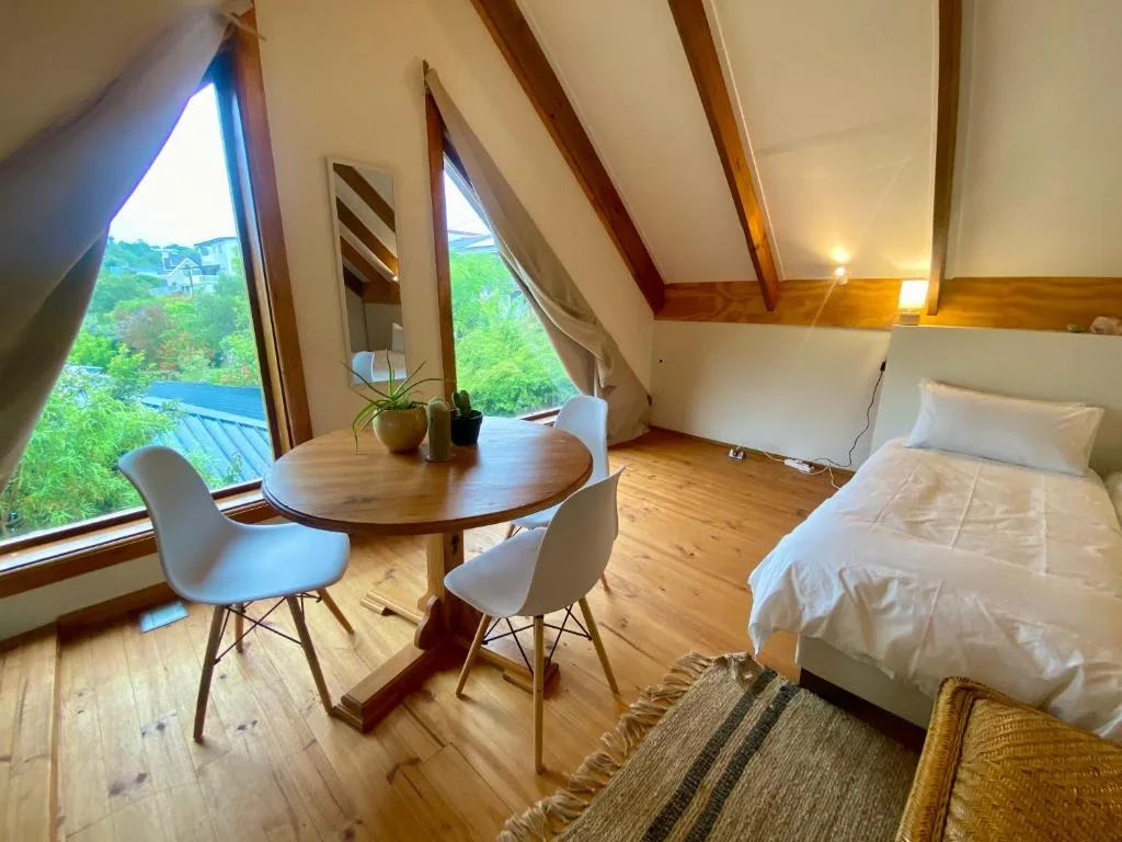 Bright loft bedroom with wooden beams, bed, round table, and scenic garden views