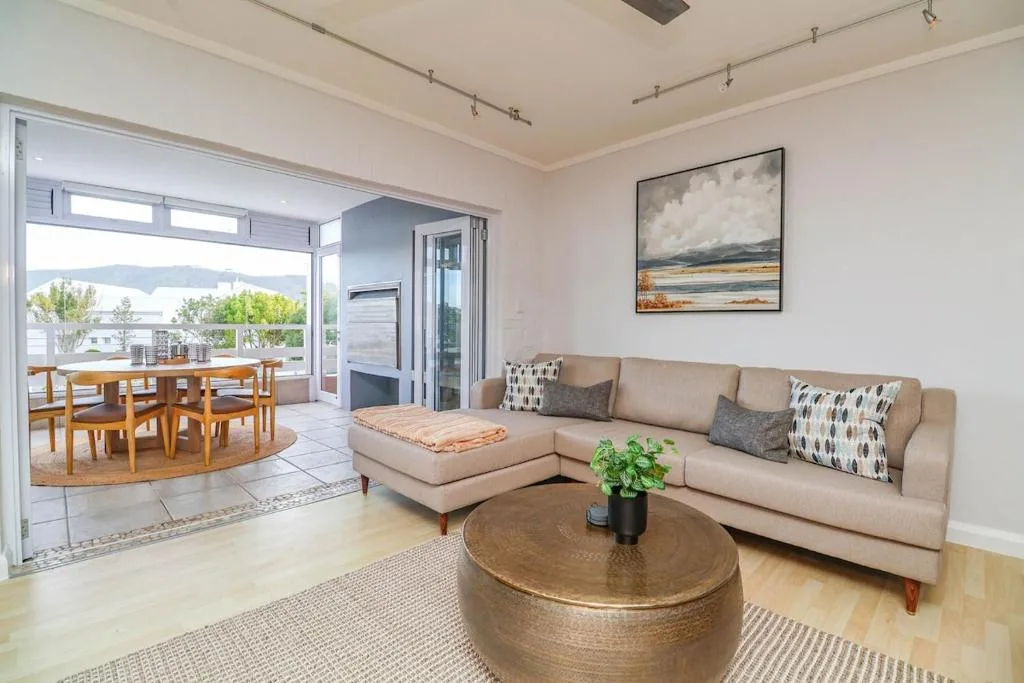 Spacious lounge with beige sectional sofa, round wooden table, and scenic lagoon views