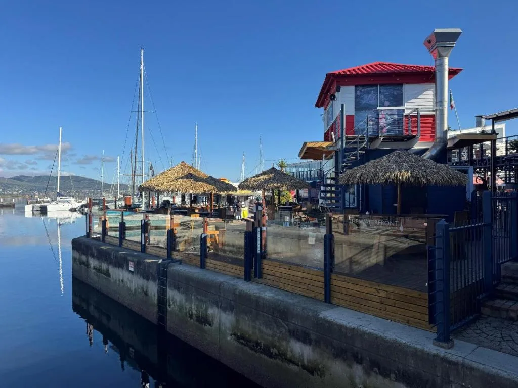 Waterfront marina with sailboats, thatched-roof structures, and mountain backdrop