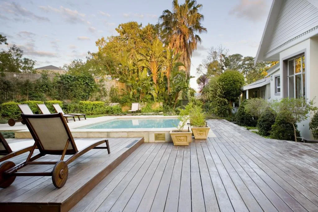 Swimming pool surrounded by wooden deck, loungers, and lush garden