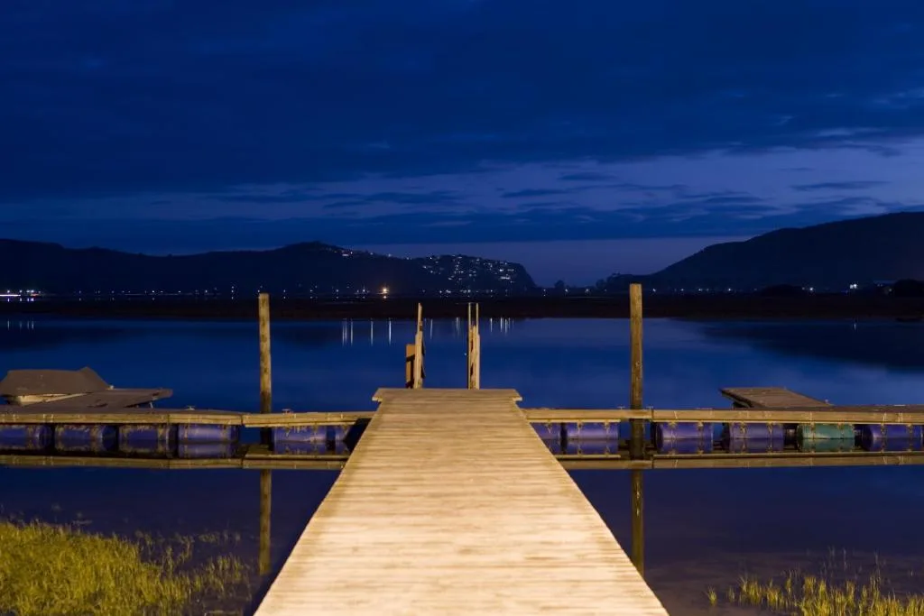 Illuminated wooden jetty extending into calm lagoon at dusk with mountains