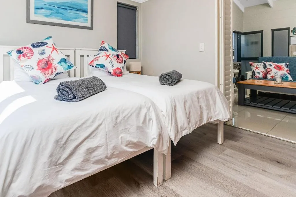 Bright bedroom with white bed, colorful floral pillows, rolled towels, and ensuite bathroom visible
