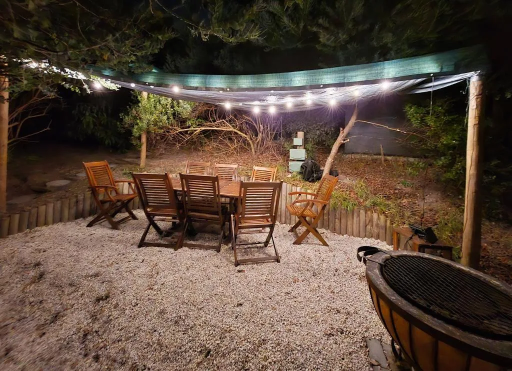 Lit outdoor entertainment area with wooden chairs and braai setup at night