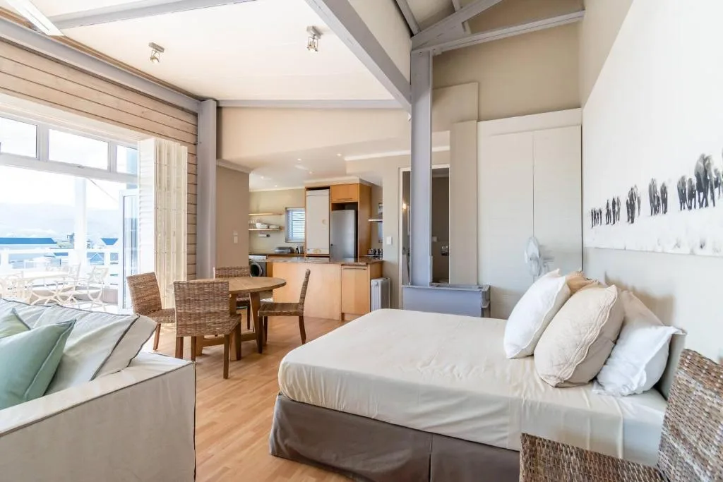 Bright bedroom with queen bed, ocean view through windows, open-plan kitchen beyond