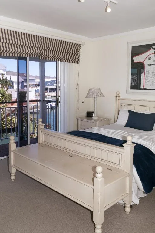 Bright bedroom with white wooden bed, striped roman blinds, waterfront balcony view