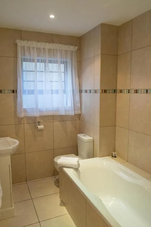 Cream-tiled bathroom with bath, toilet, and window with sheer curtains