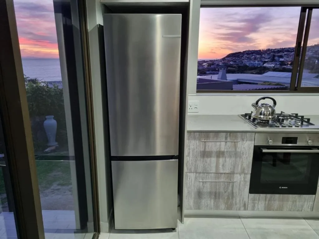 Modern kitchen with stainless steel fridge, gas cooktop, and sunset ocean views through windows