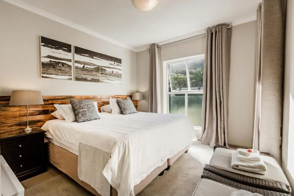 Bright bedroom with white bedding, wooden headboard, and lagoon views through large windows
