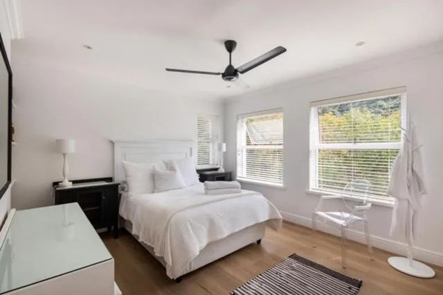 Bright modern bedroom with white bedding, ceiling fan, and garden views