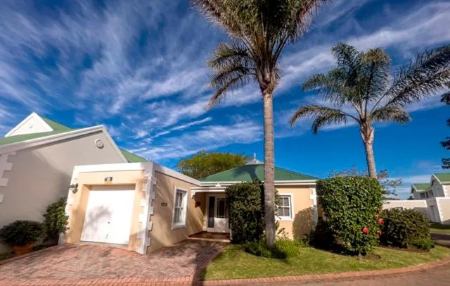 Single-story cream cottage with palm trees and brick driveway under blue sky