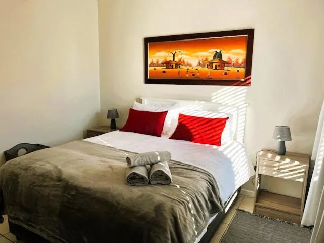 Bright bedroom with made bed, red pillows, and framed artwork above headboard