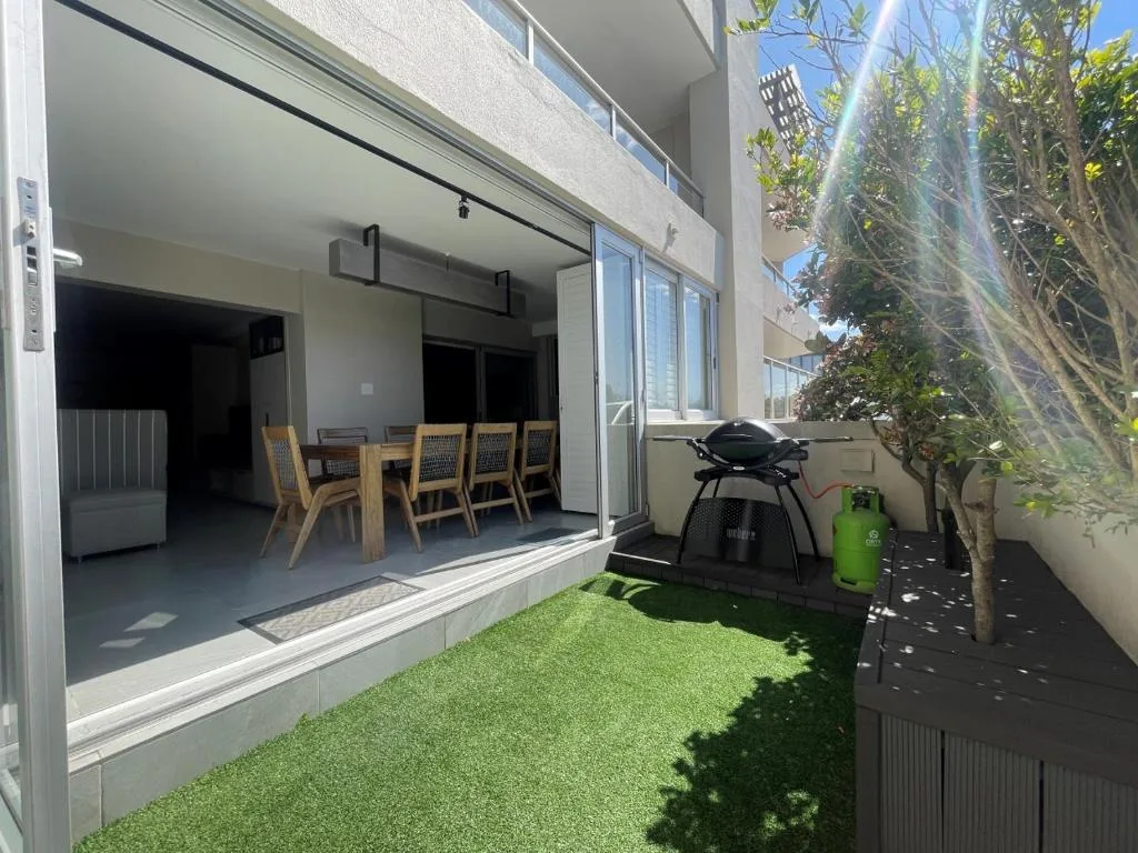Compact outdoor patio with artificial turf, dining chairs, and braai grill